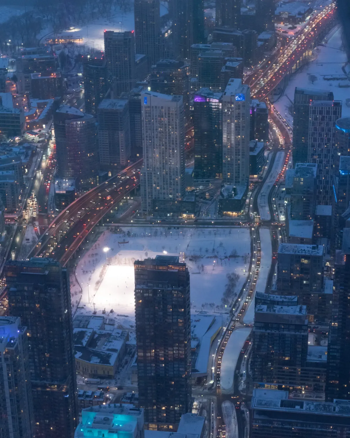 A wintry blue-hour cityscape from above, with car lights threading between snow dusted highrises and a small lit ice rink in a park below.
